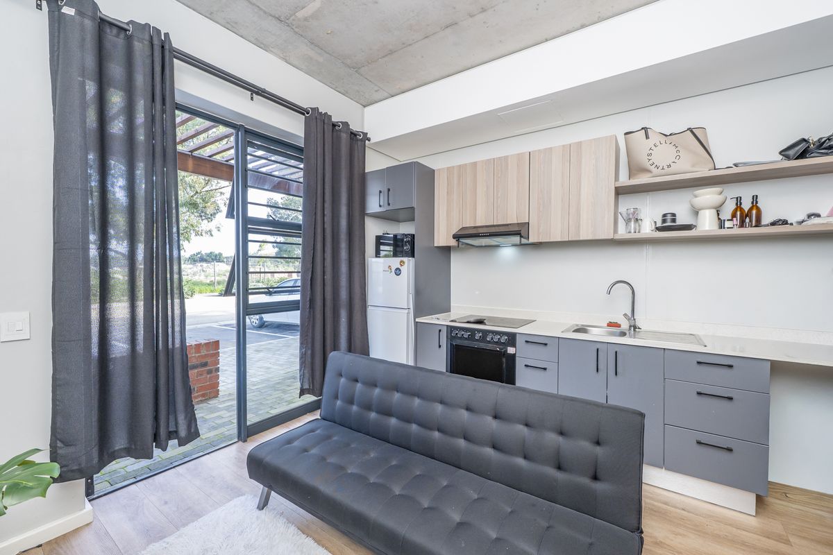 Modern open-plan studio apartment featuring a compact kitchenette with built-in appliances, exposed concrete ceiling, and direct access to an outdoor patio area.