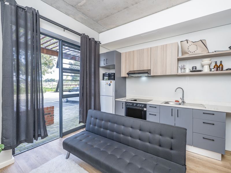 Modern open-plan studio apartment featuring a compact kitchenette with built-in appliances, exposed concrete ceiling, and direct access to an outdoor patio area.