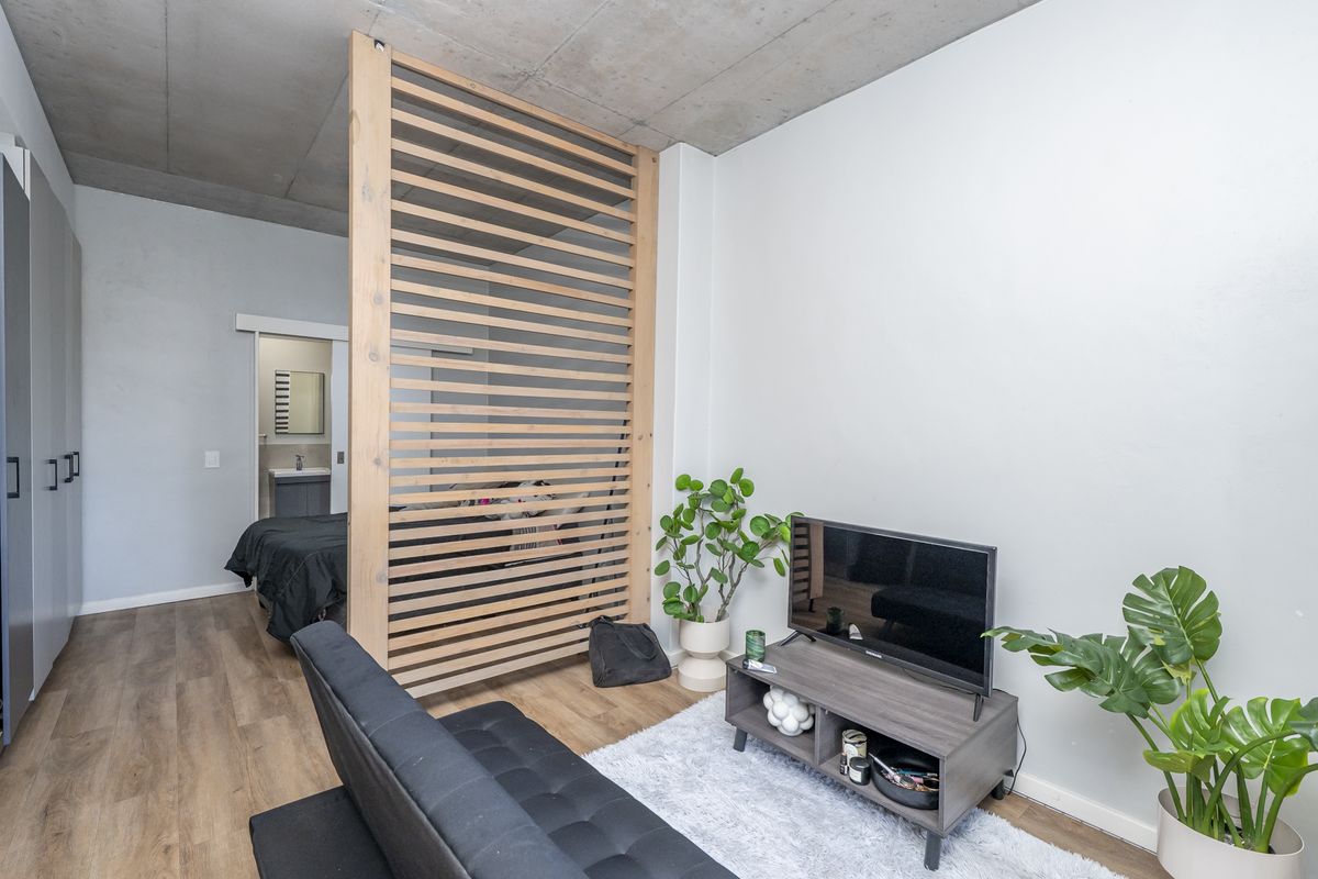 Modern bedroom featuring an exposed concrete ceiling, timber slat room divider, and laminate flooring, with an adjacent en-suite bathroom visible through a sliding door.