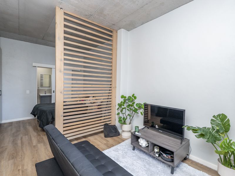 Modern bedroom featuring an exposed concrete ceiling, timber slat room divider, and laminate flooring, with an adjacent en-suite bathroom visible through a sliding door.