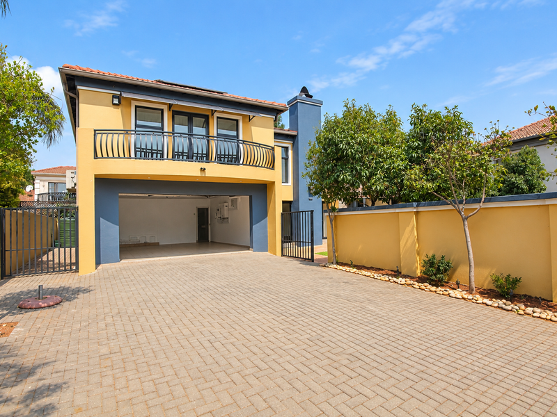 A well-maintained front exterior featuring a paved driveway, a double-story facade with a balcony, and secure perimeter walling.