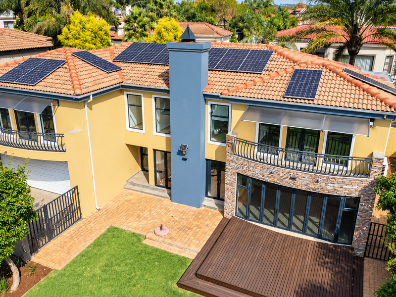 Aerial view of the rear elevation featuring a tiled roof with solar panels, stone-clad accents, timber decking, and a paved patio area.