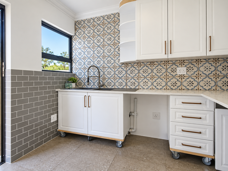 A modern, well-appointed scullery featuring white cabinetry, stone countertops, patterned tile backsplash, and dedicated plumbing connections for laundry or secondary kitchen use.