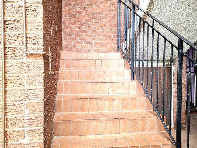 Exterior staircase with tiled steps and metal handrail leading to the apartment entrance, featuring exposed brick walls and external plumbing.