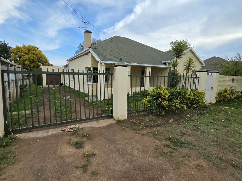 Front exterior view of a single-story house featuring a metal security gate, corrugated roof, and a large front garden area.