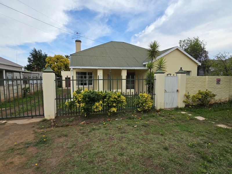 Front view of a single-story house featuring a corrugated metal roof, secure perimeter fencing, and a gated entrance with a front garden.