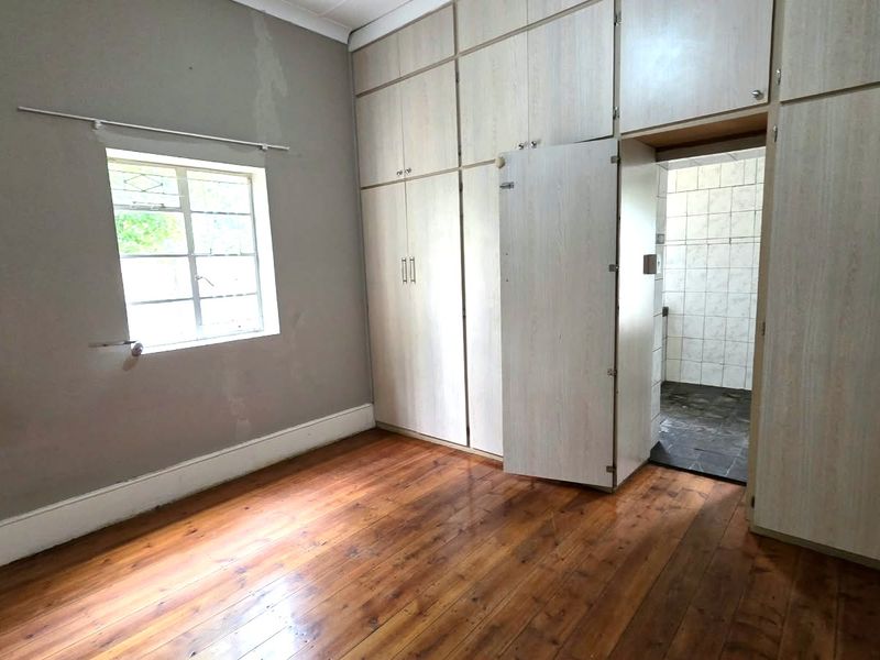 A vacant bedroom featuring original timber flooring, extensive built-in cabinetry, and a window with security bars, leading into an adjacent tiled bathroom area.