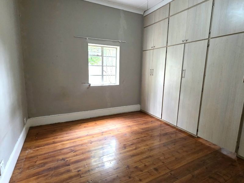 An empty bedroom featuring original timber flooring, built-in cupboards, and a window with security bars, requiring cosmetic updates to the wall finishes.
