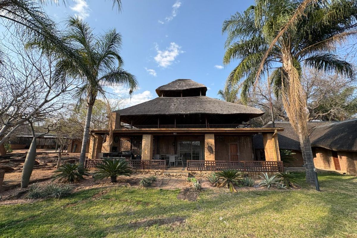A spacious rear exterior view of a farm-style residence featuring a traditional thatch roof, stone masonry, and a large covered patio area.