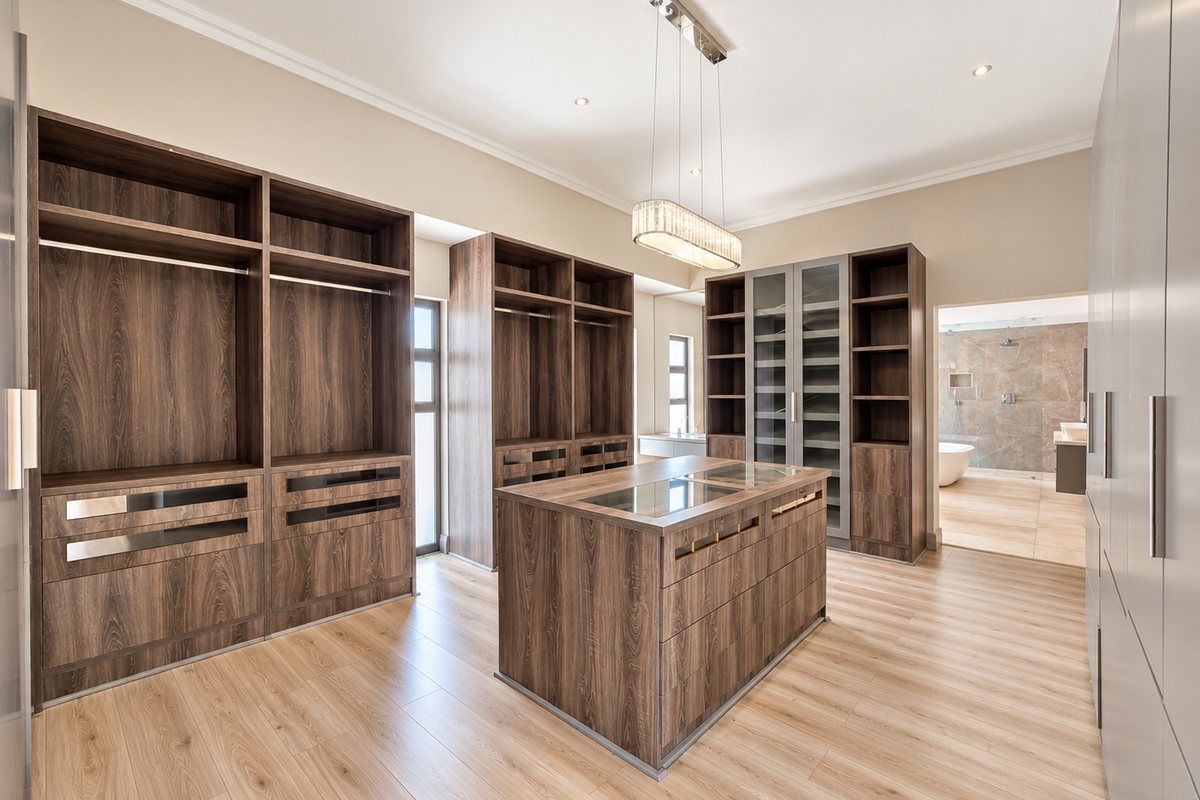 Spacious walk-in dressing room featuring extensive built-in wooden cabinetry, a central storage island, and modern lighting, with direct access to an en-suite bathroom.