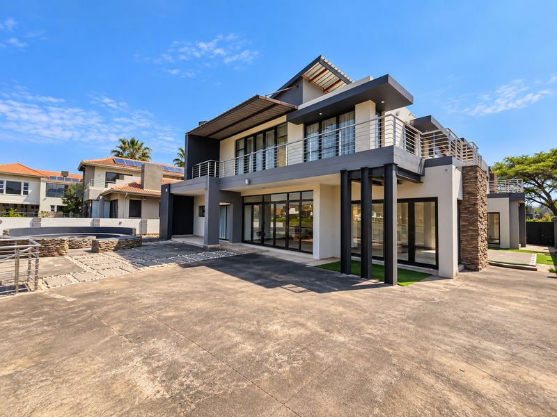 Modern multi-story residence featuring stone cladding, expansive glass sliding doors, a spacious paved patio, and a balcony with metal railings.