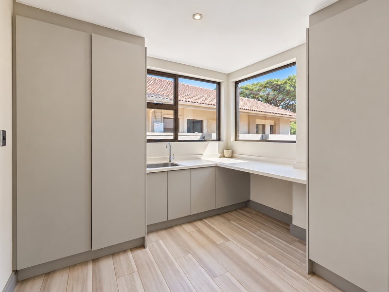 A modern, clean scullery featuring neutral built-in cabinetry, stone countertops, a stainless steel sink, and large windows providing ample natural light.