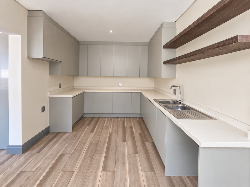 A modern, empty kitchen featuring sleek grey cabinetry, stone countertops, a stainless steel sink, and floating timber shelving with wood-look laminate flooring.