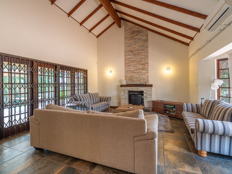 Spacious living room featuring a vaulted ceiling with exposed timber beams, a stone-clad fireplace, and tiled flooring with access to the patio.
