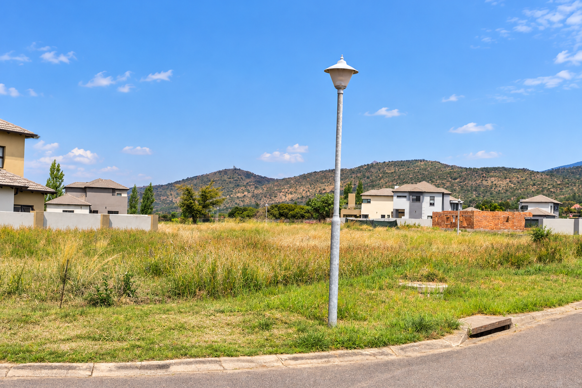 A vacant residential plot featuring established street infrastructure, including a lamp post, curb, and drainage, set against a scenic mountain backdrop.