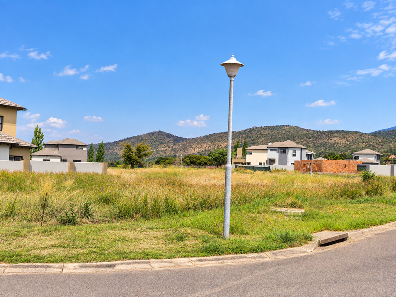 A vacant residential plot featuring established street infrastructure, including a lamp post, curb, and drainage, set against a scenic mountain backdrop.