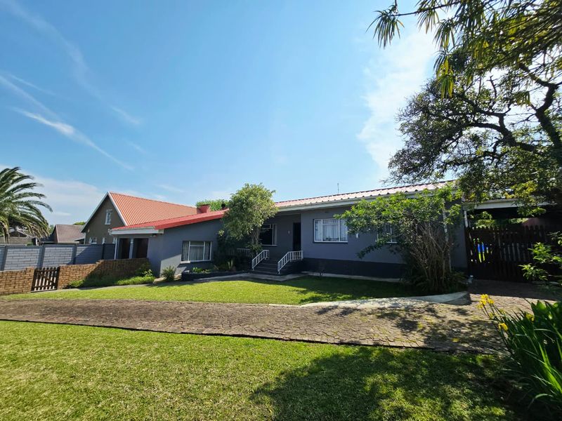 Front view of a single-story house featuring a metal roof, paved driveway, well-maintained lawn, and a brick boundary wall with a timber gate.