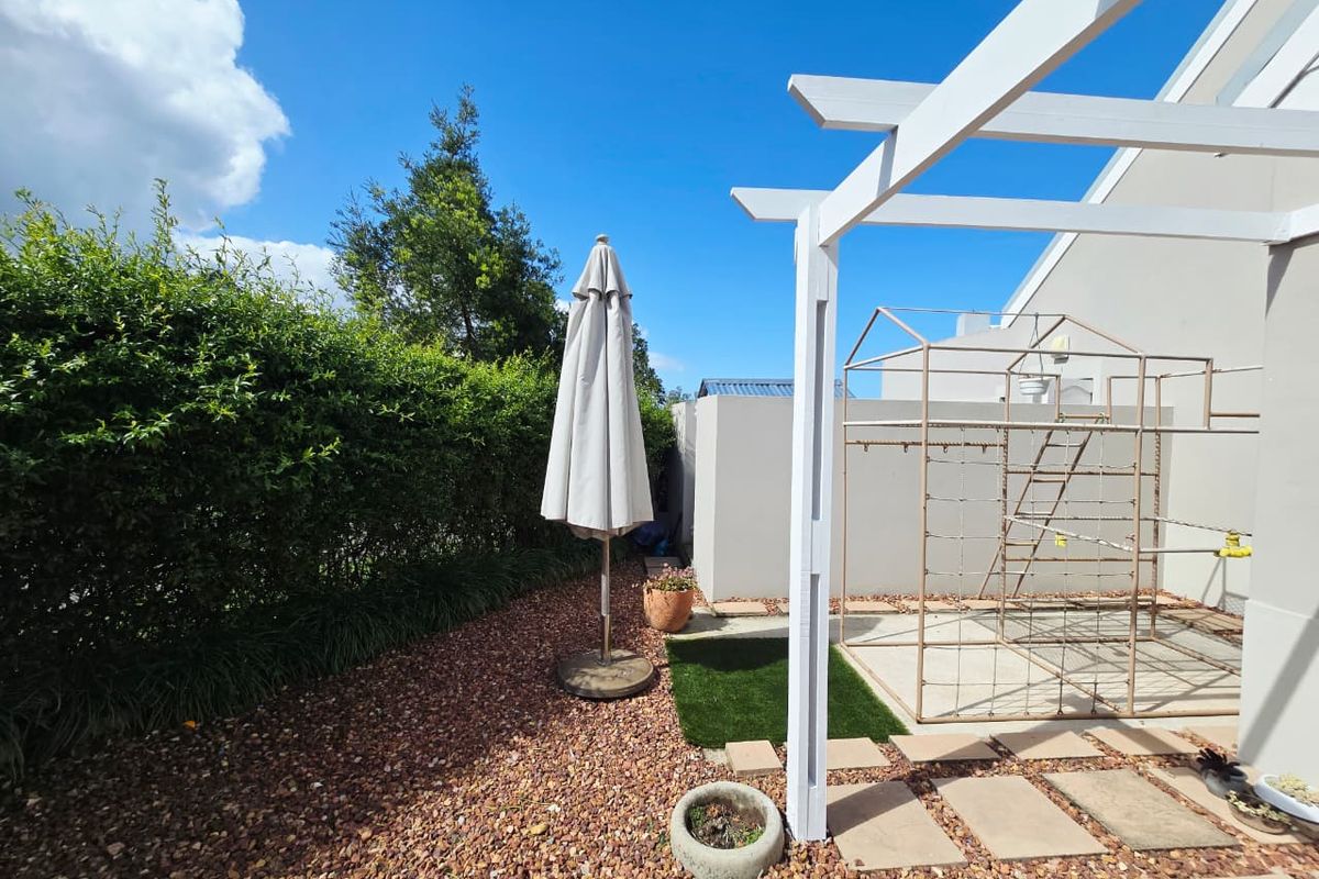 A private rear garden area featuring a white timber pergola, paved pathways, gravel landscaping, and established boundary hedging.
