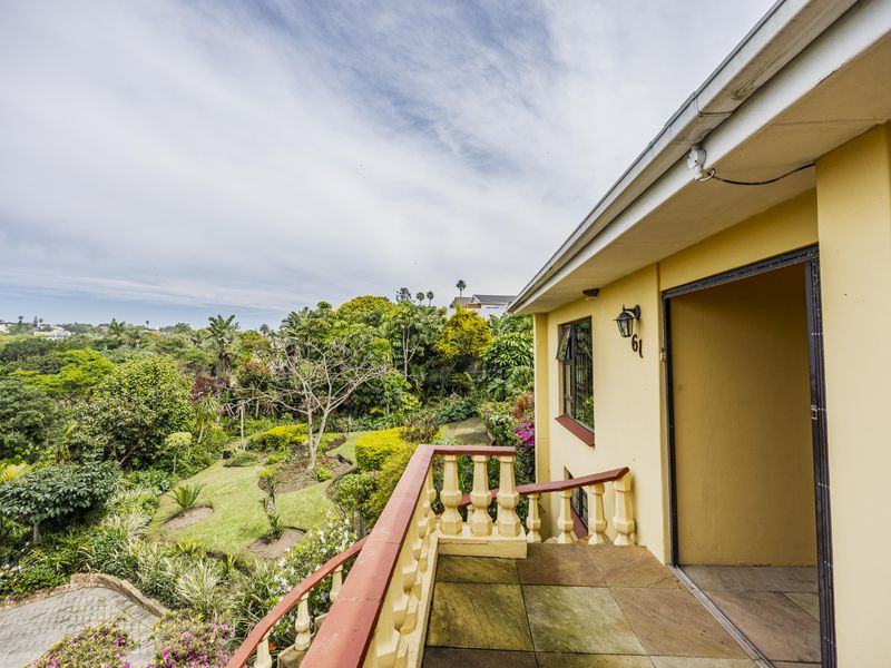 A covered balcony featuring a balustrade and paved flooring, offering an elevated view over the lush garden and surrounding landscape.
