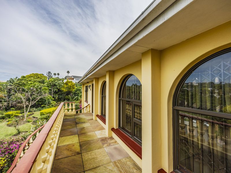 A spacious covered balcony featuring arched windows, stone tile flooring, and a decorative balustrade overlooking the lush garden.