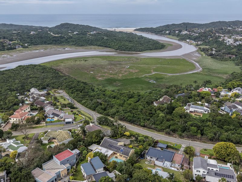 An elevated aerial view showcasing the coastal residential estate, surrounding natural vegetation, and the nearby estuary and ocean coastline.