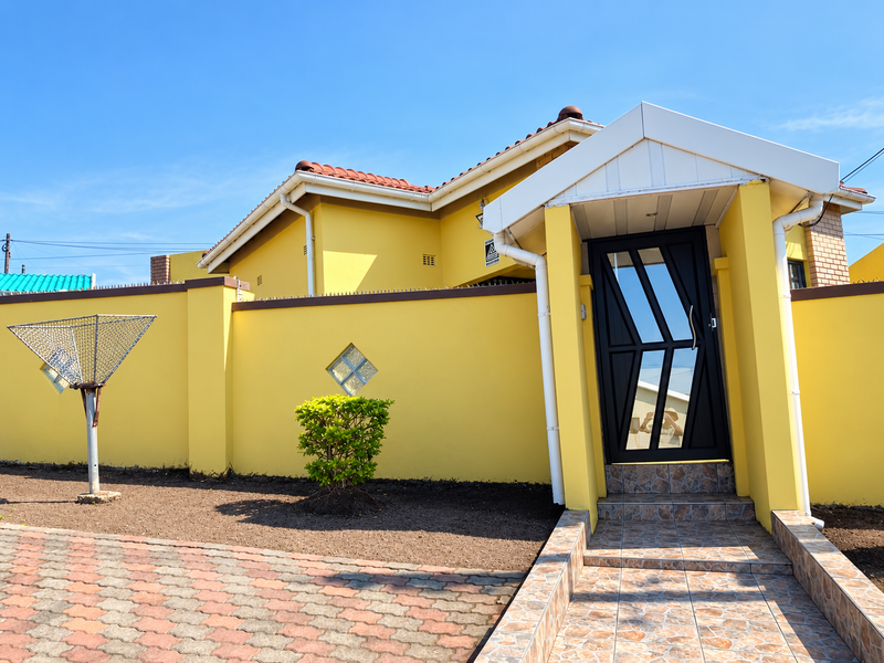 Front entrance of a yellow house featuring a tiled porch, modern security gate, and a paved driveway with perimeter walling.