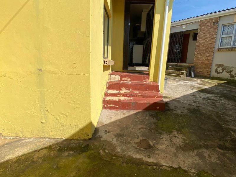 Rear exterior view showing a yellow painted wall, tiled entrance steps, and concrete paving with visible signs of weathering and surface growth.