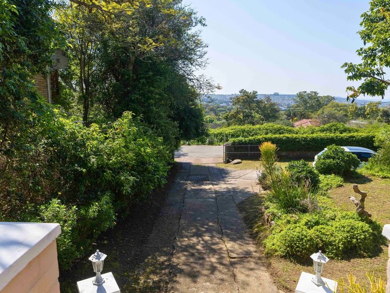 Front view of the property featuring a concrete driveway, established garden, and gated entrance with distant neighborhood views.