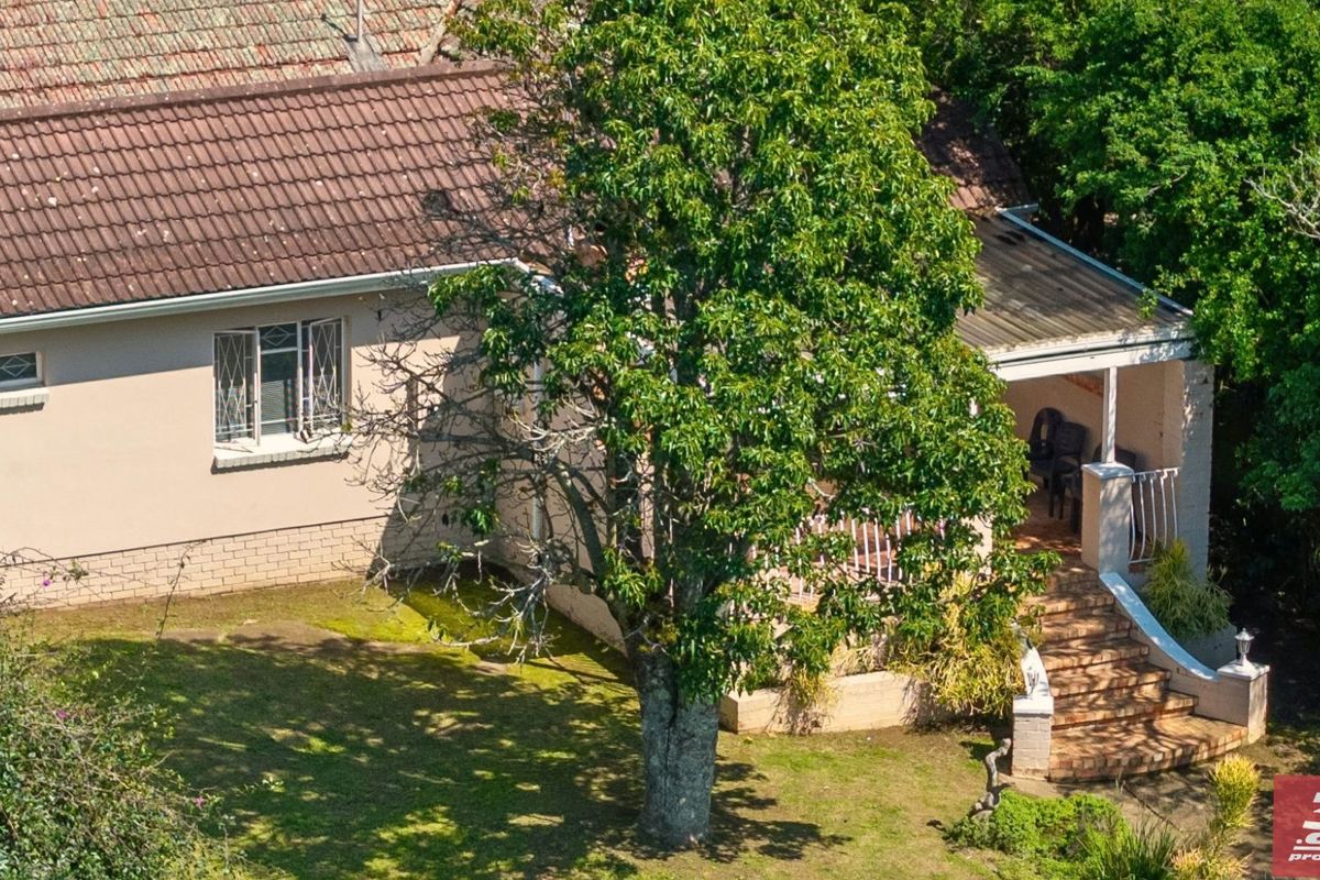 An elevated view of the rear exterior featuring a tiled roof, a covered patio area with brick stairs, and established garden surroundings.
