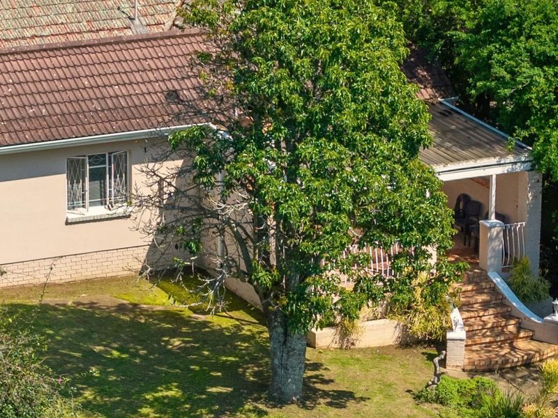 An elevated view of the rear exterior featuring a tiled roof, a covered patio area with brick stairs, and established garden surroundings.