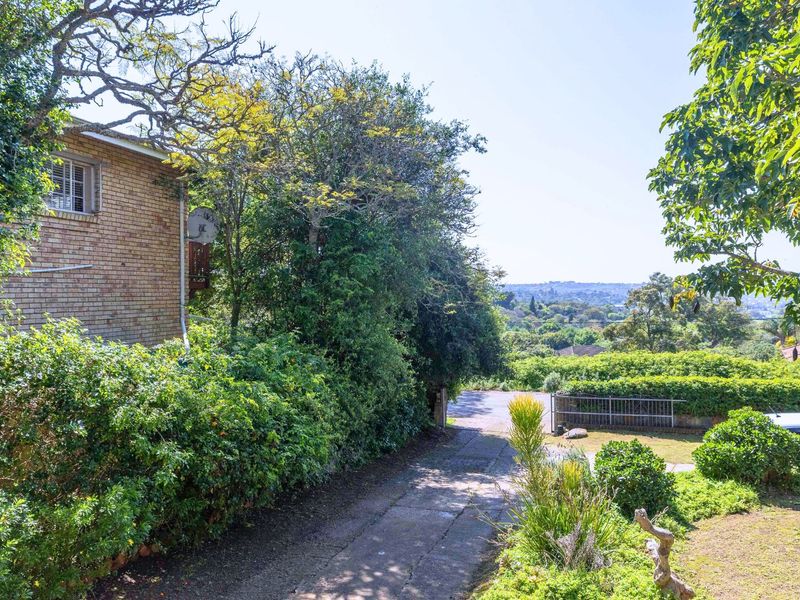 A view of the property's front exterior, featuring a brick facade, a paved driveway, and established garden landscaping with a scenic outlook.