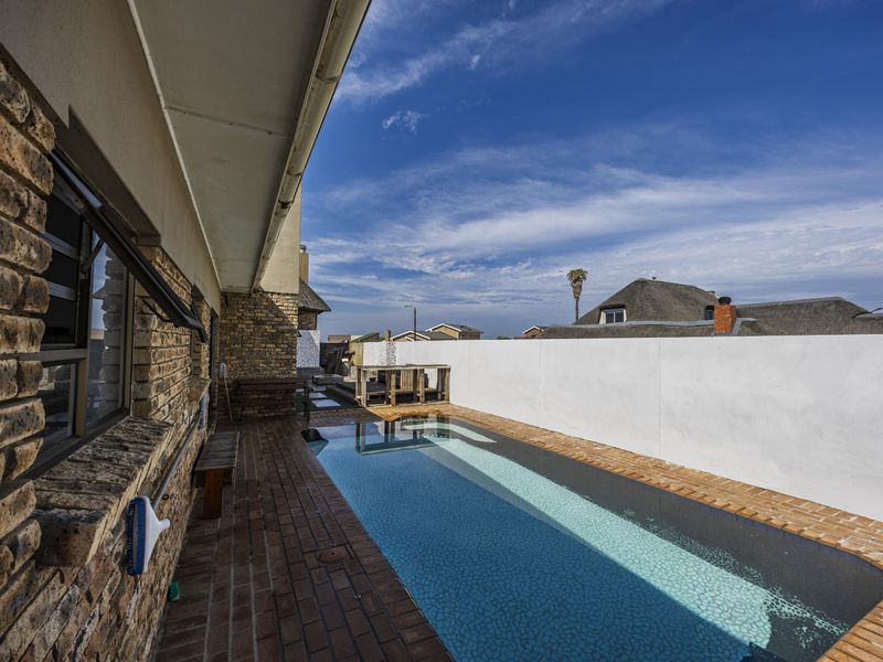 A narrow lap-style swimming pool set within a brick-paved courtyard, featuring a covered patio area and a high boundary wall for privacy.
