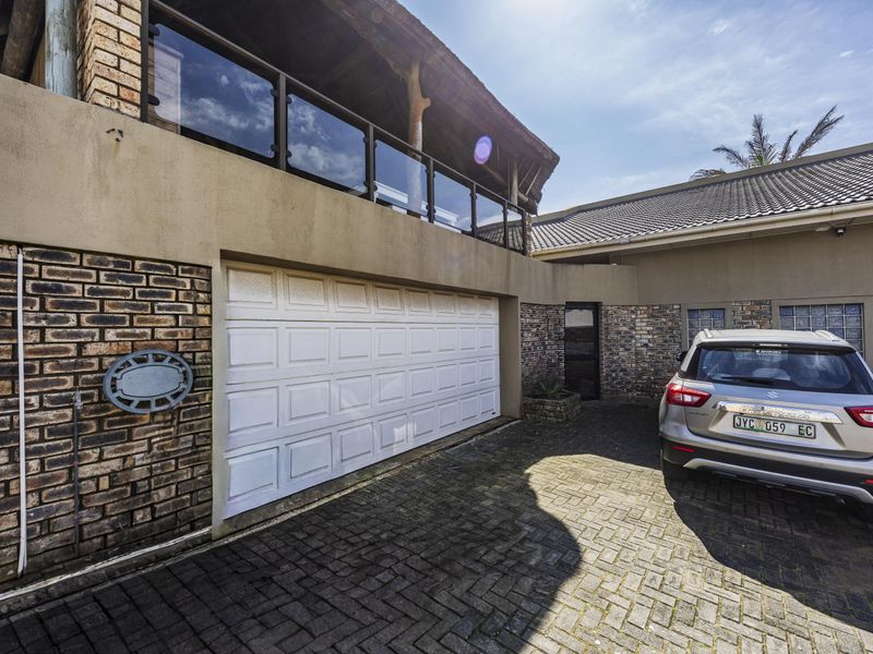 Front exterior view of the property featuring a double garage, paved driveway, and an upper-level balcony with a glass balustrade and thatched roof.