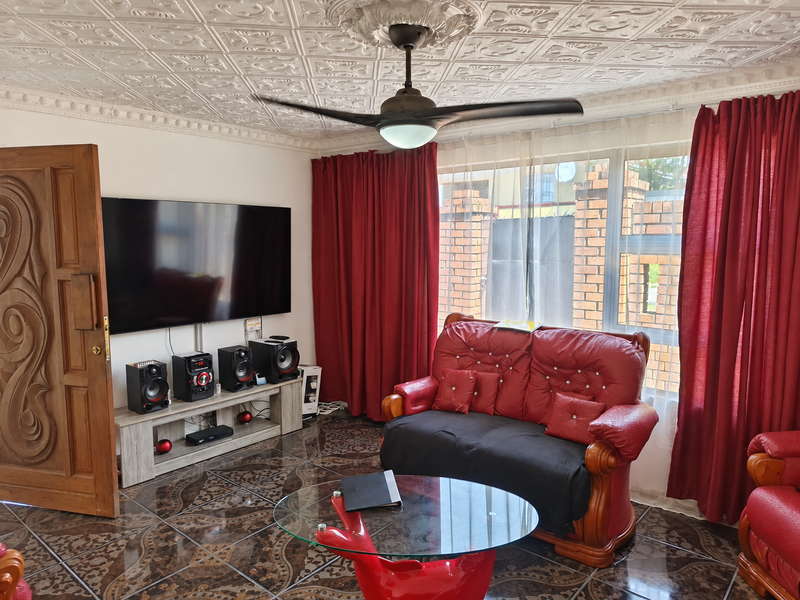A living room featuring decorative ceiling tiles, a ceiling fan, and dark patterned floor tiles, with large windows providing natural light.