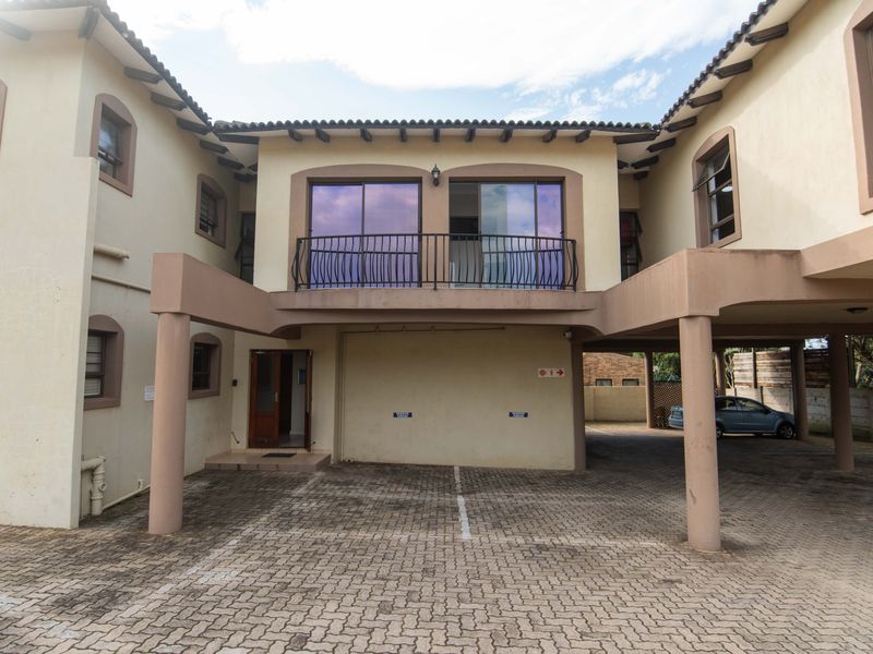 Front elevation of a townhouse complex featuring a paved driveway, covered parking bays, and an upper-level balcony with wrought iron railings.