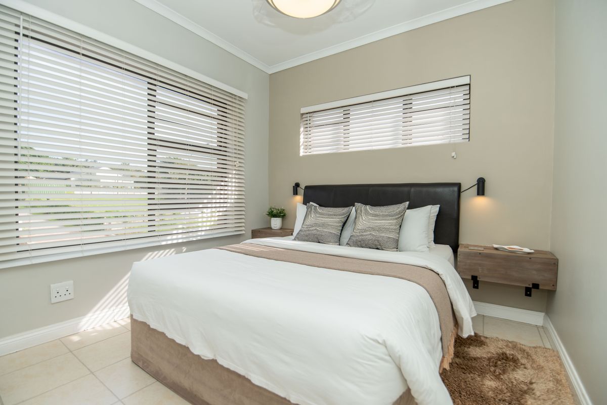 A well-maintained bedroom featuring tiled flooring, dual windows with venetian blinds, and modern wall-mounted bedside lighting with floating nightstands.