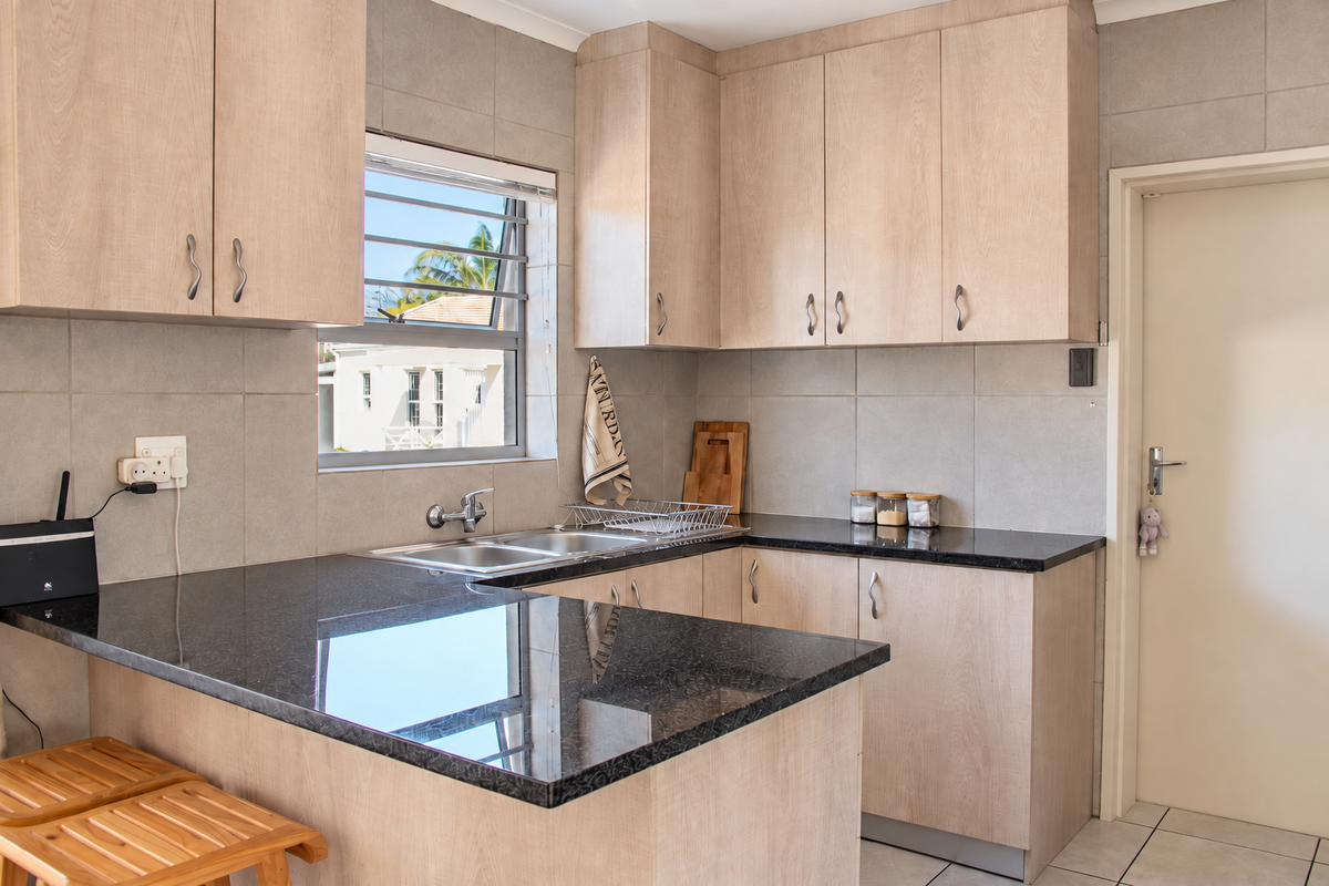 A functional kitchen featuring light wood-finish cabinetry, dark granite countertops, a double basin sink, and neutral tiled wall finishes.