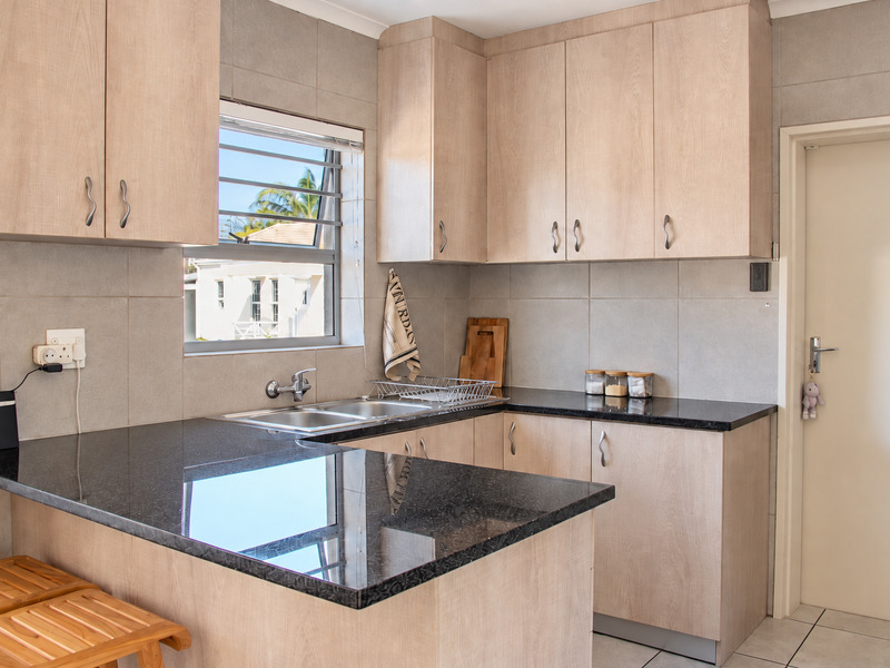 A functional kitchen featuring light wood-finish cabinetry, dark granite countertops, a double basin sink, and neutral tiled wall finishes.