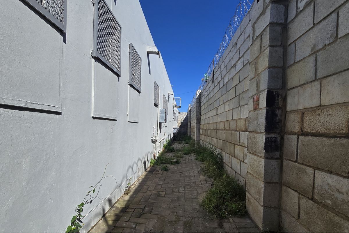 A narrow side passage featuring a paved walkway, perimeter wall with security razor wire, and exterior wall with security grilles.