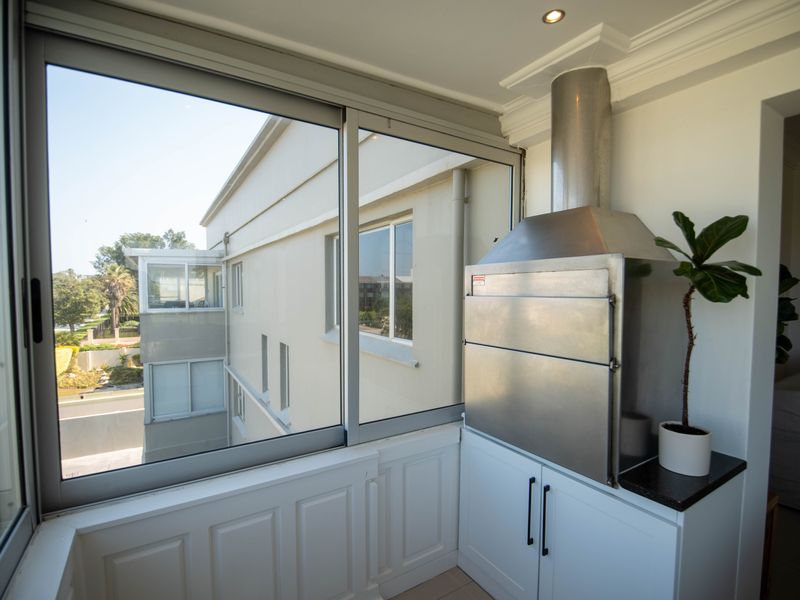 Enclosed balcony featuring a built-in stainless steel braai, modern cabinetry, granite countertop, and large sliding glass windows with views of the surrounding complex.