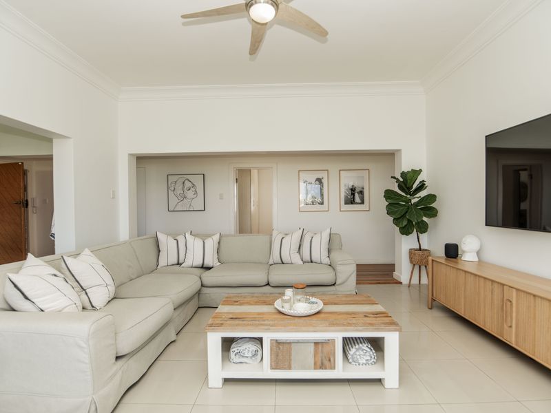 A bright and well-maintained living room featuring neutral tiled flooring, a ceiling fan, and clean white walls with decorative crown molding.