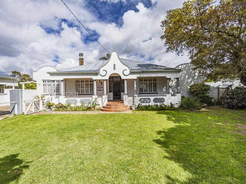 Charming single-story residence featuring a classic gabled facade, covered front porch with columns, and a spacious front garden with gated driveway access.