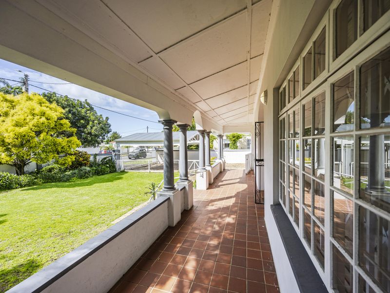 A covered veranda featuring terracotta floor tiles, decorative columns, and large multi-pane windows overlooking a well-maintained garden.