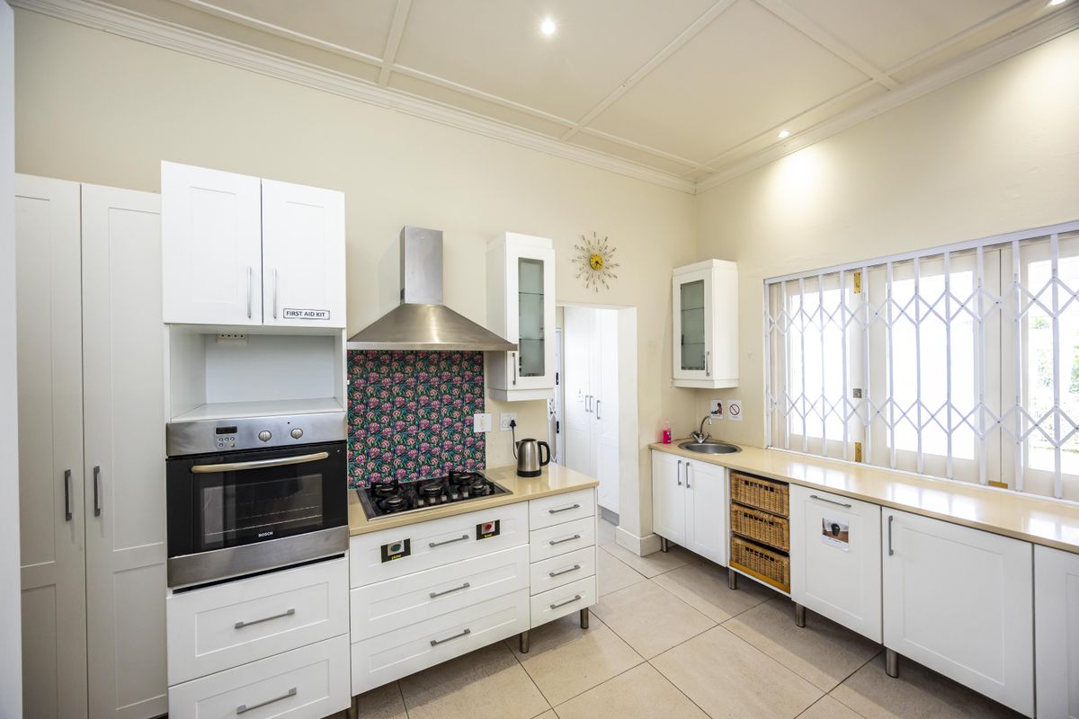 A functional kitchen featuring white cabinetry, built-in oven, gas hob with extractor, tiled flooring, and a stainless steel sink with security gate on the window.