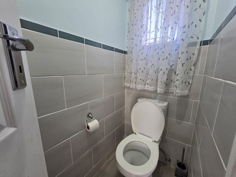 A compact bathroom featuring neutral wall tiling, a standard toilet, and a window with privacy curtain.