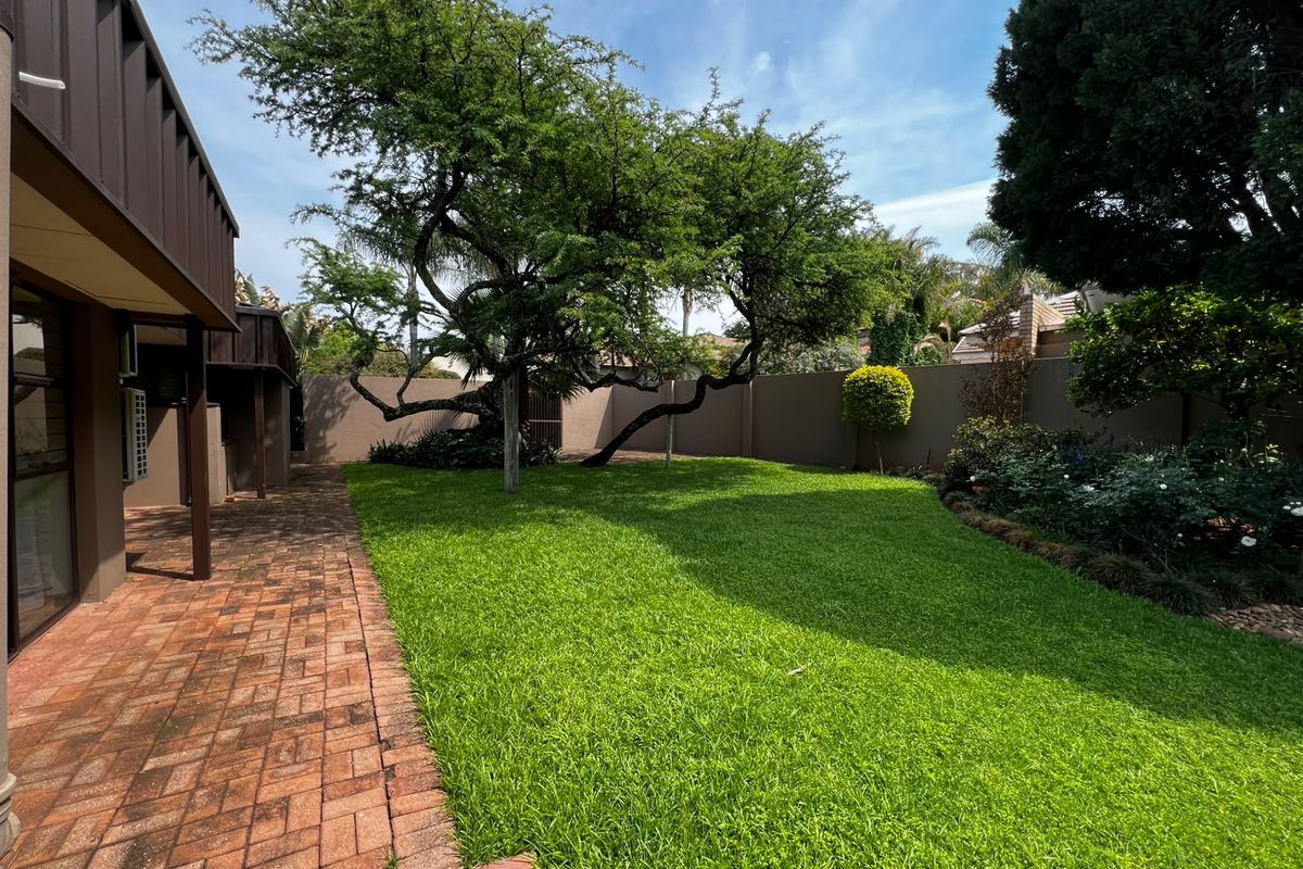 A well-maintained rear garden featuring a brick-paved patio area, lush green lawn, and established trees enclosed by a solid boundary wall.