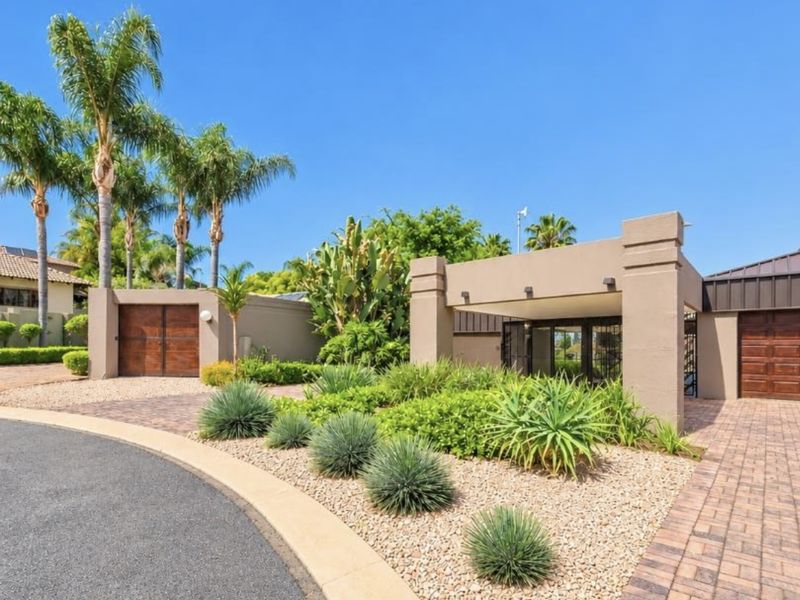 A well-maintained front exterior featuring a paved driveway, covered entrance portico, wooden garage doors, and established landscaped garden beds.