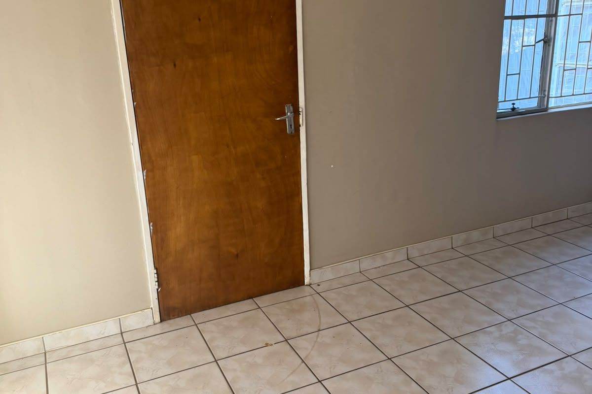 An empty bedroom featuring ceramic floor tiles, a wooden door, and a window fitted with security bars.