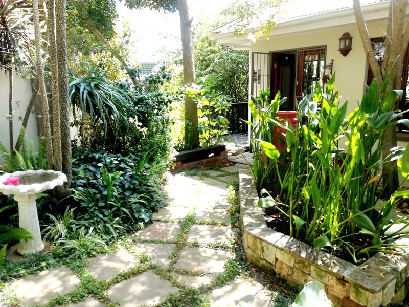 A lush, established garden featuring a stone-paved pathway, a raised stone planter, and mature greenery leading toward the home's exterior entrance.