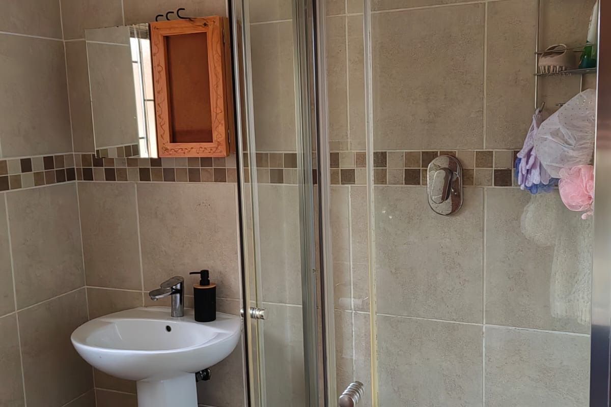A functional bathroom featuring a pedestal basin, a glass-enclosed shower with a rain shower head, and neutral ceramic wall tiling with a mosaic accent.
